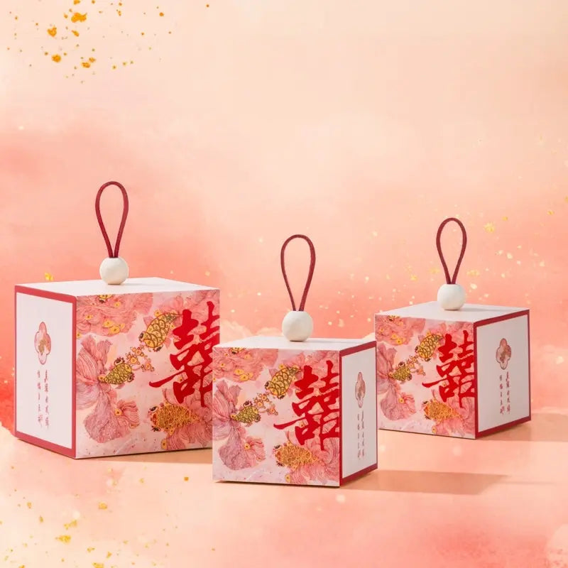 Three  square Floral & Koi Fish Double Happiness Wedding Favor Boxes with red hanging loops and white beads, printed with pink floral motifs and large red Chinese characters, arranged on a soft peach-pink background.
