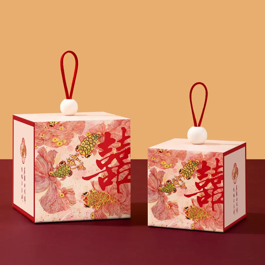 Two Floral & Koi Fish Double Happiness Wedding Favor Boxes  with red hanging loops and white spherical tops, printed with pink floral motifs, gold fish, and large red Chinese characters, displayed on a dark red surface against a warm beige background.