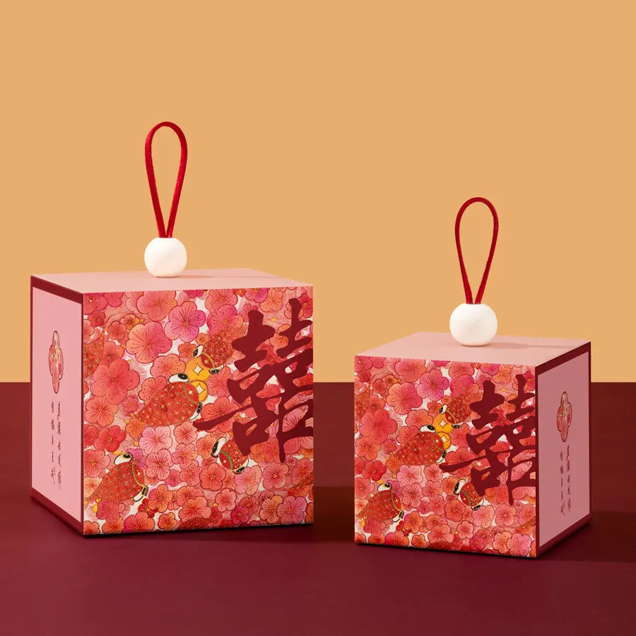 Two Floral & Koi Fish Double Happiness Wedding Favor Boxes s with pink floral patterns, large Chinese double happiness symbols, and red hanging loops with white beads, displayed on a dark red surface against a warm beige background.