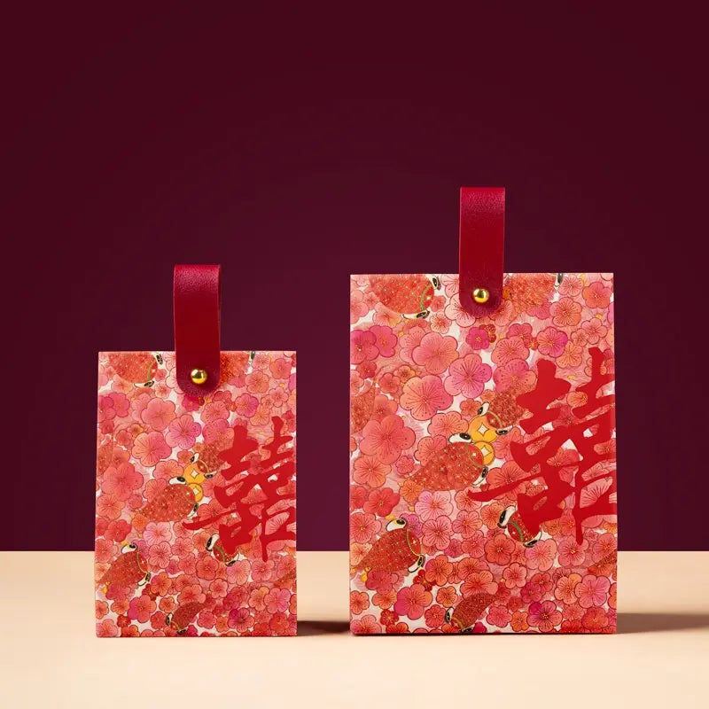 Two red and pink Floral & Koi Fish Double Happiness Wedding Favor Boxes matching red ribbon handles and gold-tone metal loops, displayed on a light beige surface against a dark burgundy background. The boxes feature Chinese double happiness symbols and a small bird illustration.