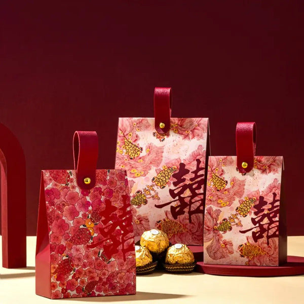 Three red and pink Floral & Koi Fish Double Happiness Wedding Favor Boxes With Handle with gold Chinese double-happiness symbols and gold foil-wrapped chocolates, arranged on a beige surface against a dark red background.