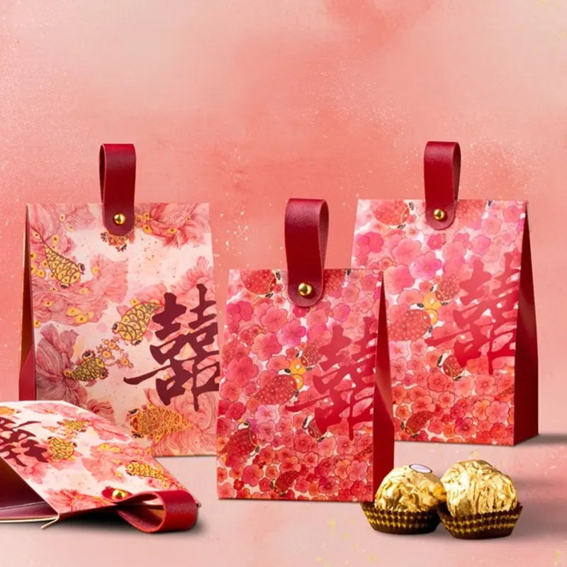 Three red and pink Floral & Koi Fish Double Happiness Wedding Favor Boxes With Handle with gold Chinese double happiness characters, shown with a matching envelope and two wrapped chocolates on a pink background.