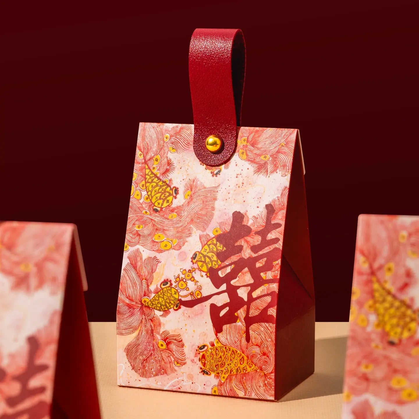 A red and pink Floral & Koi Fish Double Happiness Wedding Favor Boxes With Handle with a gold-tone snap strap and a large Chinese double happiness symbol, shown on a tabletop against a dark red background.