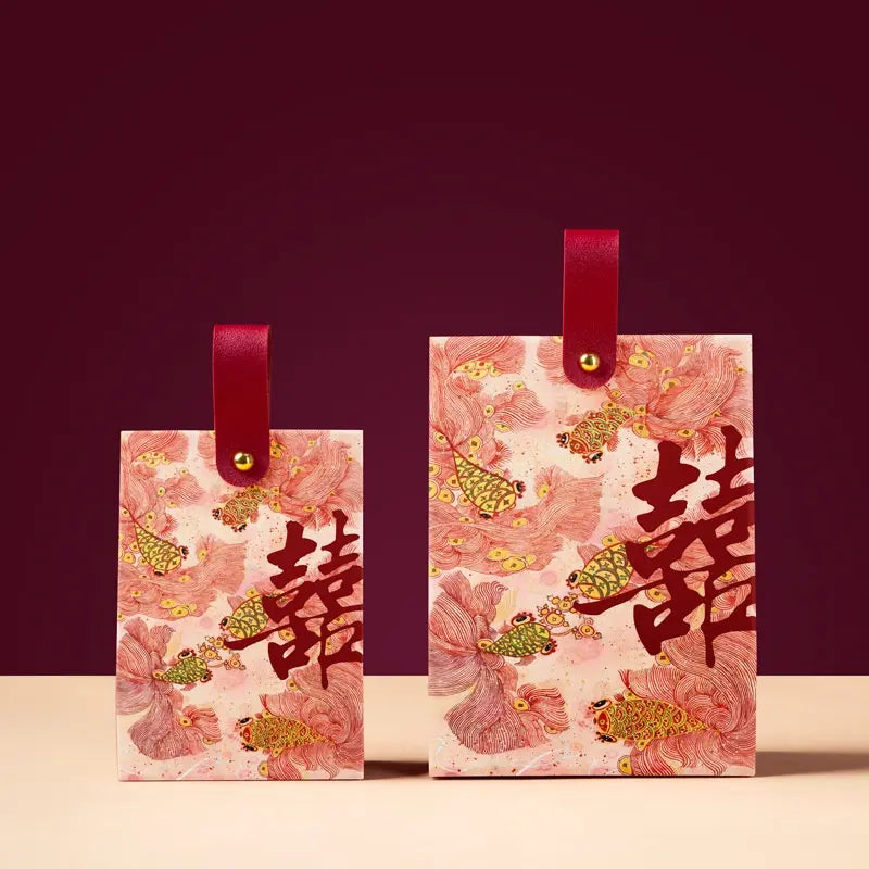 Two Floral & Koi Fish Double Happiness Wedding Favor Boxes   with red leather loops and gold-tone hardware, printed with pink floral motifs, gold fish, and large red Chinese double-happiness symbols, shown on a beige surface against a dark burgundy background.