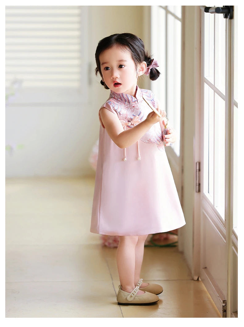Young girl in a pink Girls' "Good Fortune" Traditional Sleeveless A-Line Jacquard Qipao dress standing in a room with light-colored walls and a door.