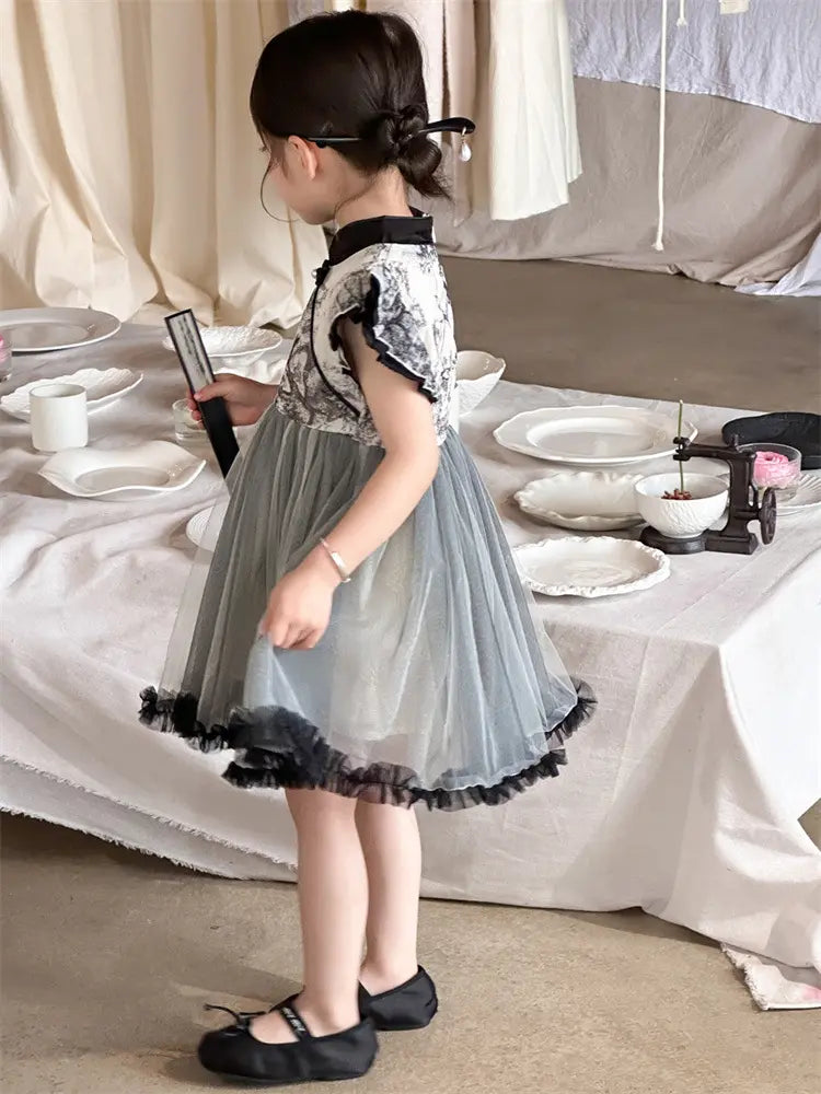 A young girl wearing a short-sleeved gray Girls Traditional Ink Painting Sleeveless Layered Tulle Qipao Dress  with a black lace bodice and black ruffled hem, standing in a styled room with a table covered in white linens and dishes.