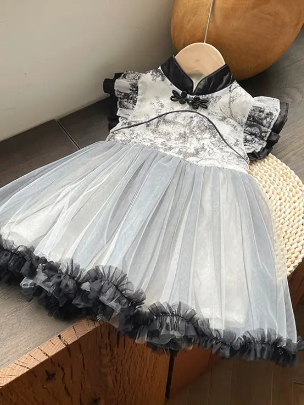 A toddler girl's dress displayed on a wooden hanger, featuring a black and white Girls Traditional Ink Painting Sleeveless Layered Tulle Qipao Dress with a mandarin collar and frog button, a sheer white tulle skirt, and a black ruffled hem.