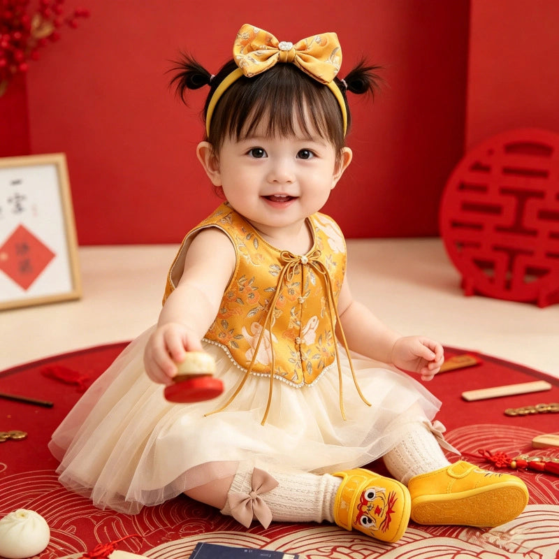 A smiling toddler girl sitting on a red patterned rug in a festive indoor setting. She wears a Golden Brocade Vest & Tulle Chinese Baby Princess Dress   with a large yellow bow headband, white socks, and yellow shoes.