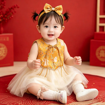 A baby girl sitting on a red patterned rug wearing Golden Brocade Vest & Tulle Chinese Baby Princess Dress , a yellow bow headband, and white socks, with red decorative boxes in the background.