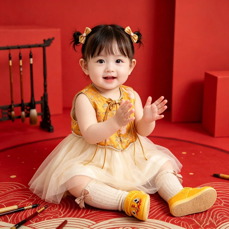 A toddler girl sits on a red patterned floor wearing a yellow and cream traditional Chinese-style dress with a tulle skirt, cream knee-high socks, and yellow shoes. She has dark hair in two buns with yellow bows.