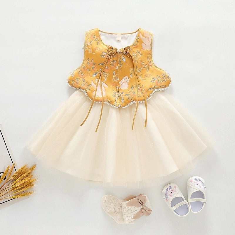 A baby girl's  Golden Brocade Vest & Tulle Chinese Baby Princess Dress  featuring a yellow embroidered vest with a bow tie and a cream tulle tutu skirt, styled with a matching cream bow headband and white baby shoes with floral prints on a light gray background.