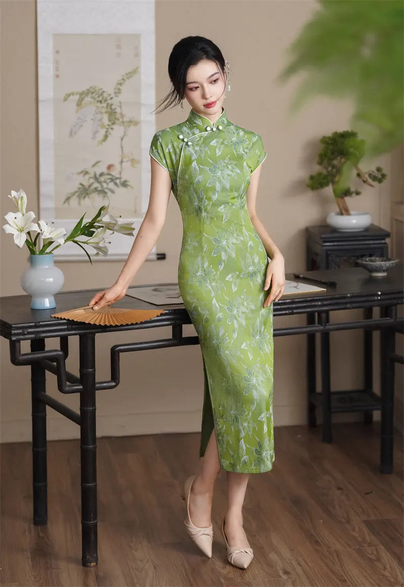 a Chinese Woman in a Green Floral  Cap Sleeve Modern Qipao Cheongsam Dressstanding in a room with a table and decorative items.
