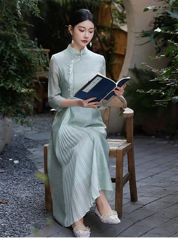 A woman sits on a wooden chair in a garden courtyard, holding an open book. She is wearing a light sage green traditional Chinese A Line qipao dress with pleated detailing and lace embroidery on the collar and sleeves. 