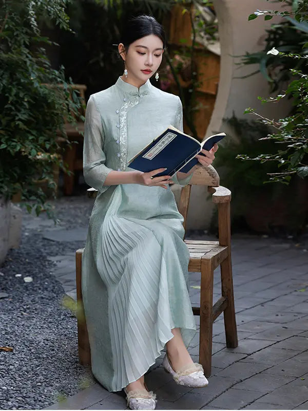 A woman sits on a wooden chair in a garden courtyard, holding an open book. She is wearing a light sage green traditional Chinese A Line qipao dress with pleated detailing and lace embroidery on the collar and sleeves. 