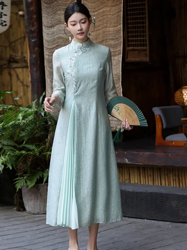 A woman wearing a light sage green long-sleeve midi a line qipao cheongsam dress with a pleated front panel and embroidered collar, holding a green folding fan in a traditional-style courtyard setting.