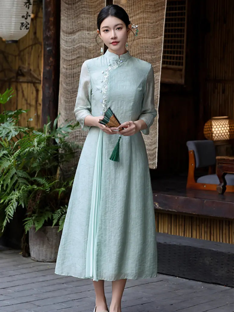 A woman wearing a light sage green traditional Chinese dress (cheongsam) with three-quarter sleeves and a high collar, standing in a rustic courtyard setting.