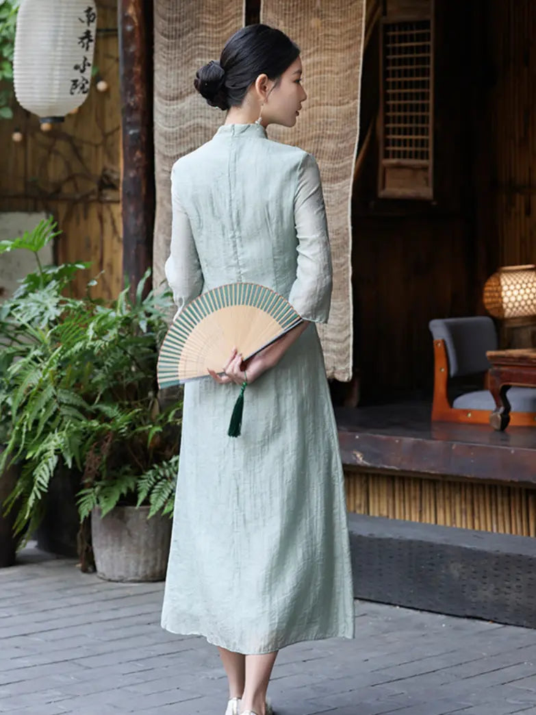 A woman stands with her back to the camera wearing a light sage green long-sleeved midi a line qipao cheongsam dress and holding a beige folding fan with green accents, set against a traditional wooden courtyard background.