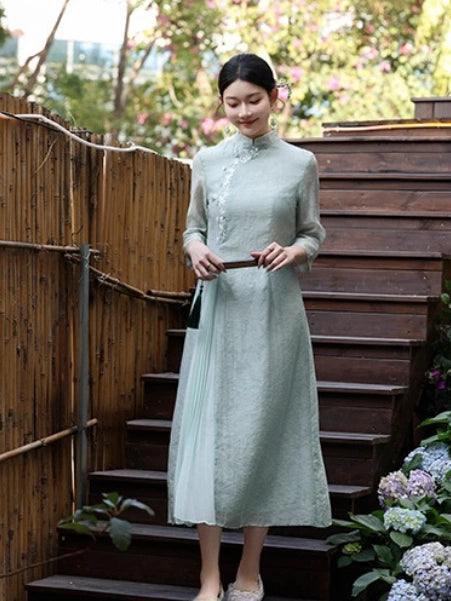 A woman wearing a light sage green midi a line qipao cheongsam dress with a high collar and long sleeves, standing on outdoor wooden steps next to a bamboo fence.