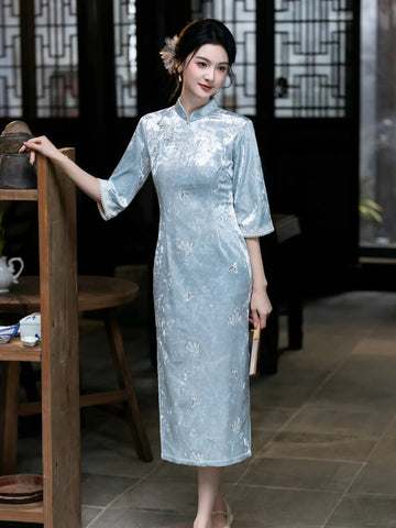 A woman wearing a light blue floral embroidered cheongsam (qipao) dress, standing in a traditional Chinese-style interior with wooden furniture and screens.