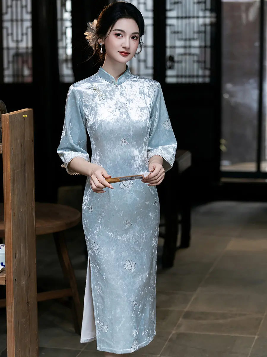 A woman wearing a light blue and silver floral-patterned cheongsam (qipao) dress, standing in a traditional Chinese courtyard setting.
