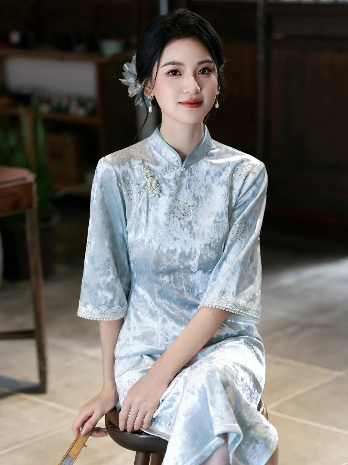 A woman wearing a light blue textured Chinese cheongsam dress, sitting on a wooden stool in a traditional indoor setting.