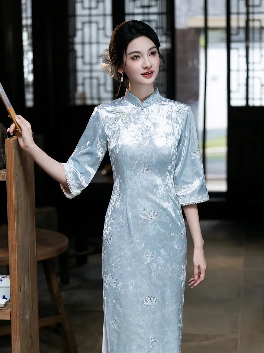 A woman wearing a light blue velvet long sleeve cheongsam with white floral embroidery, standing in a traditional Chinese-style interior.