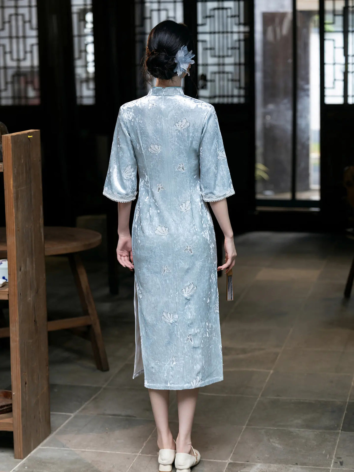 A woman viewed from behind wearing a light blue embroidered cheongsam qipao dress in a traditional Chinese-style interior with dark wood screens and tiled floors.