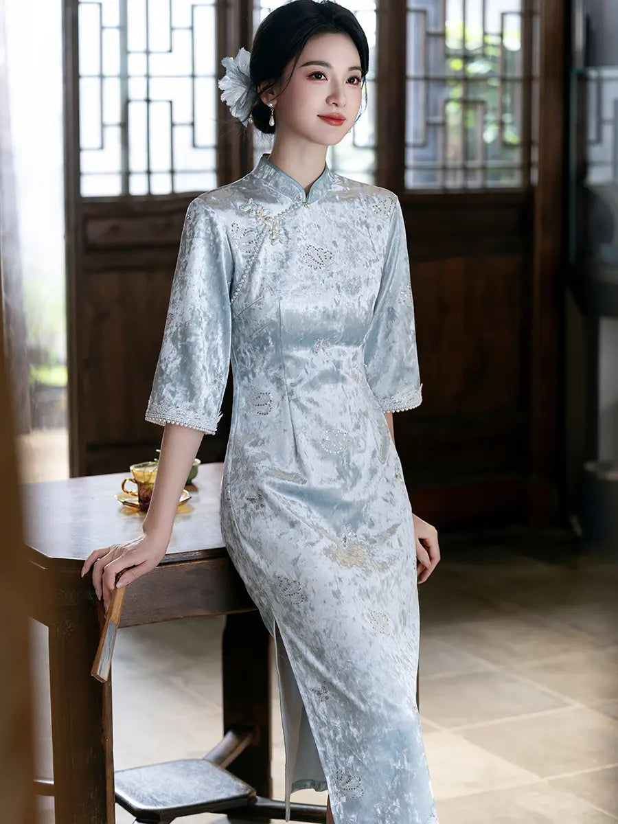 A woman wearing a light blue velvet qipao cheongsam with subtle floral embroidery, posing in a traditional Chinese courtyard next to a wooden table.