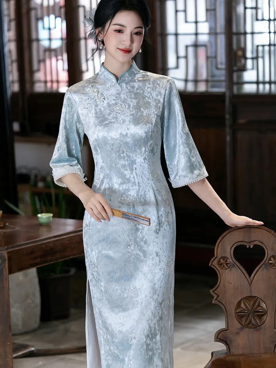 A woman wearing a light blue textured cheongsam (qipao) dress with three-quarter sleeves and a high collar, standing in a traditional Chinese room with wooden furniture and lattice windows.