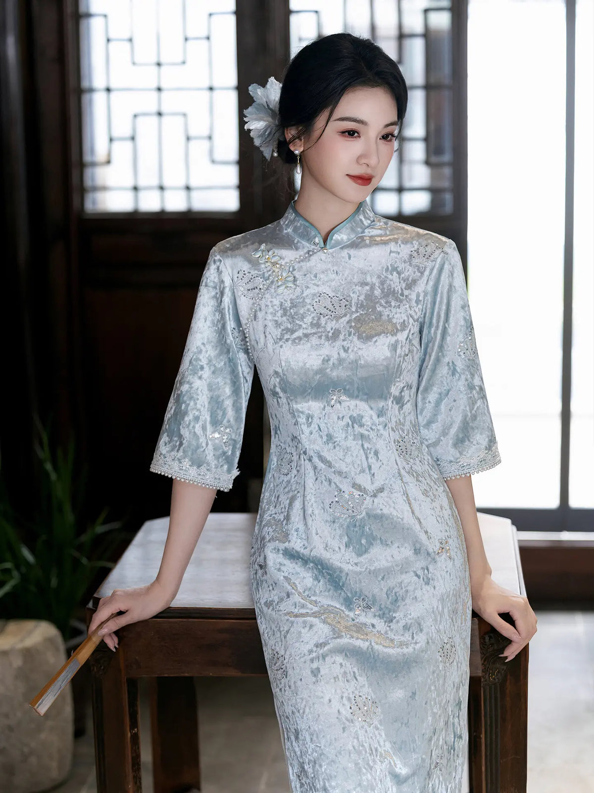 A woman wearing a light blue and silver brocade cheongsam (qipao) dress, standing in a traditional Chinese room with wooden lattice windows.