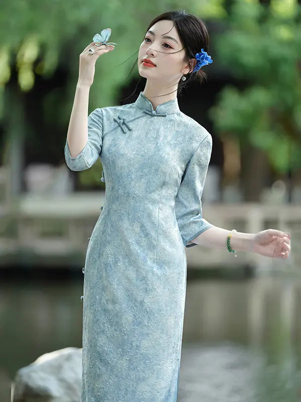 A woman wearing a light blue textured cheongsam dress stands outdoors near water and greenery, holding a butterfly near her face. The dress has a high collar, three-quarter sleeves, and side buttons.