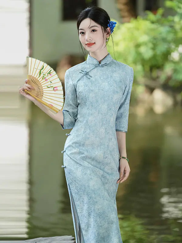 A woman wearing a light blue textured cheongsam (qipao) dress stands by a pond, holding a folding fan. The dress has a classic mandarin collar, three-quarter sleeves, and side buttons.