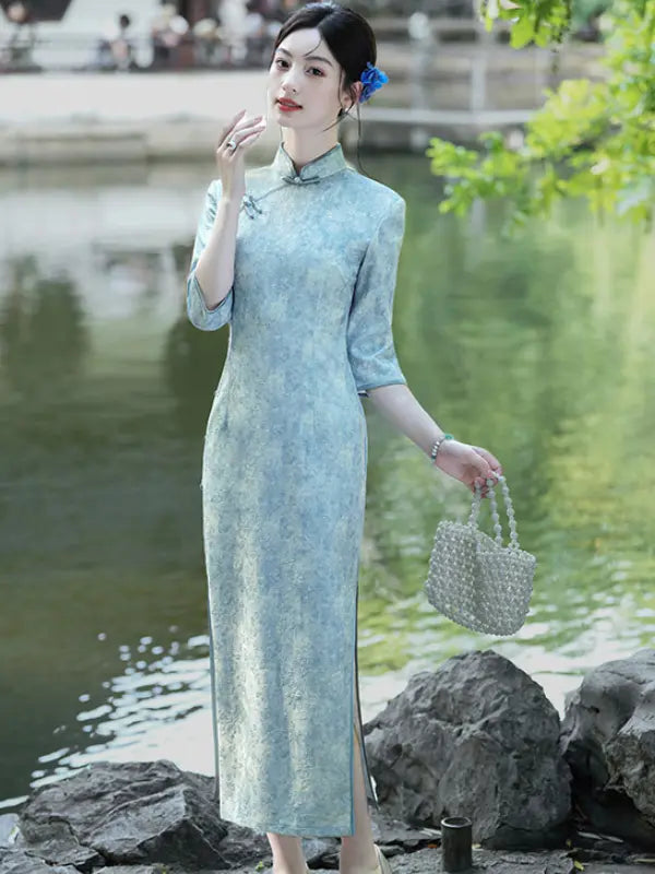 A woman wearing a light blue-green textured cheongsam dress stands by a lake, holding a woven handbag.