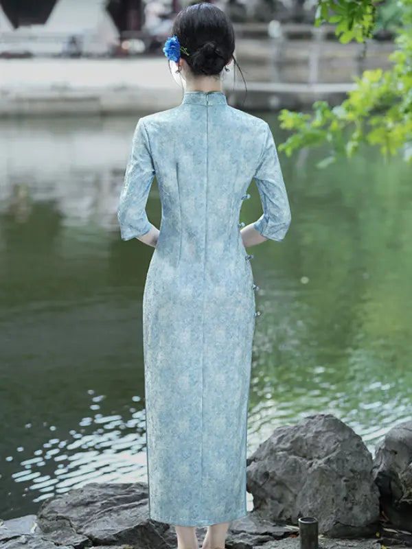 A woman stands with her back to the camera wearing a light blue textured cheongsam (qipao) dress, standing on rocks next to a calm pond with green foliage in the background.