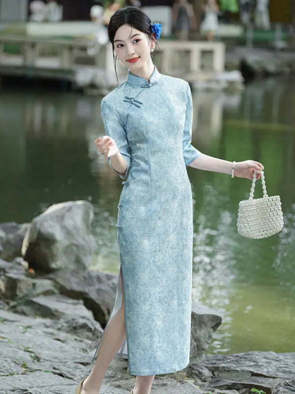 A woman wearing a light blue textured cheongsam (qipao) dress stands outdoors by a pond, holding a small woven cream handbag.