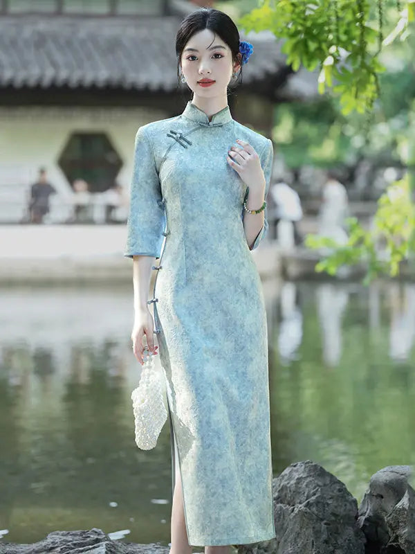 A woman wearing a light blue-green textured cheongsam (qipao) dress stands outdoors by a pond with traditional buildings and greenery in the background.