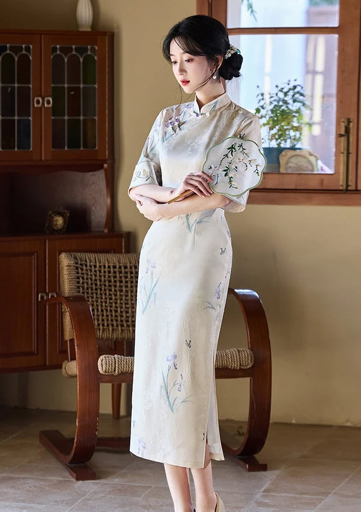 Woman in a traditional  Ivory Floral Jacquard Qipao Dress with Embroidery outfit holding a fan in a room with wooden furniture and a window.