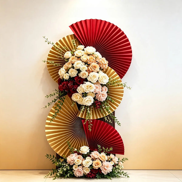 A decorative floral arrangement featuring three large pleated paper fans in red and gold, styled with cream, blush, and burgundy roses and green foliage against a warm beige wall.