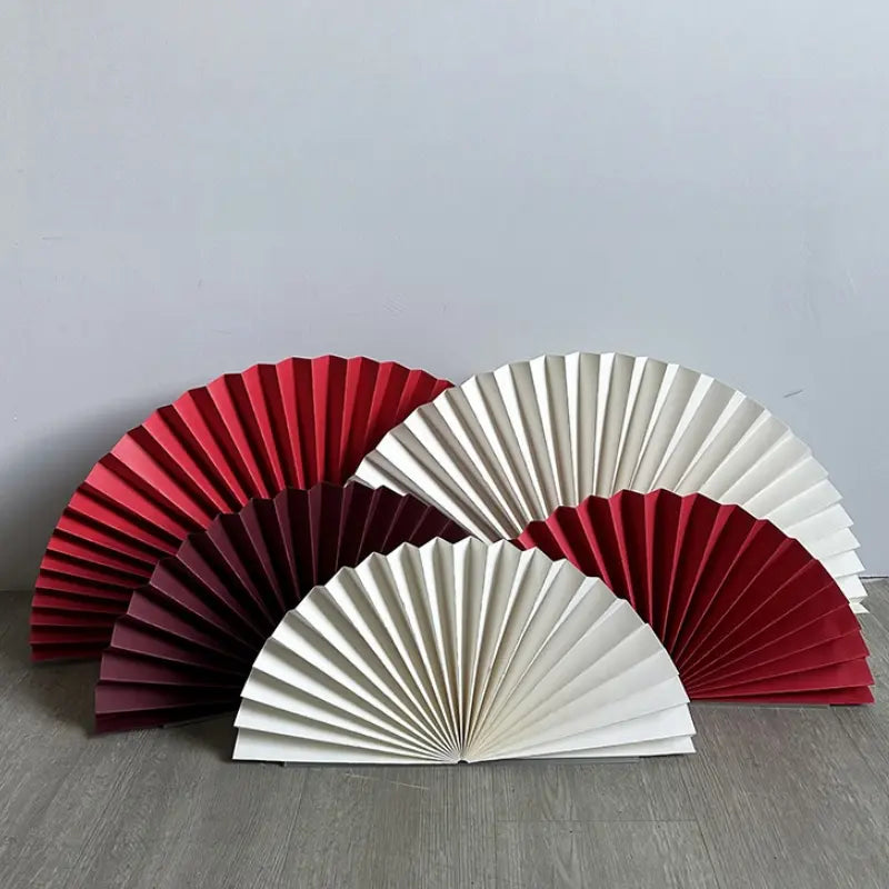 Four folding paper fans arranged on a gray floor against a light gray wall. The set includes two red fans and two cream fans, all shown open in a semi-circular shape.