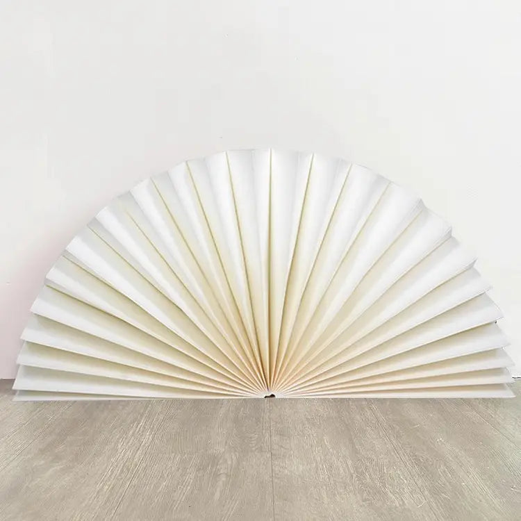A white pleated accordion-style wall fan or decorative folding screen, partially unfolded and leaning against a plain white wall on a light wood floor.