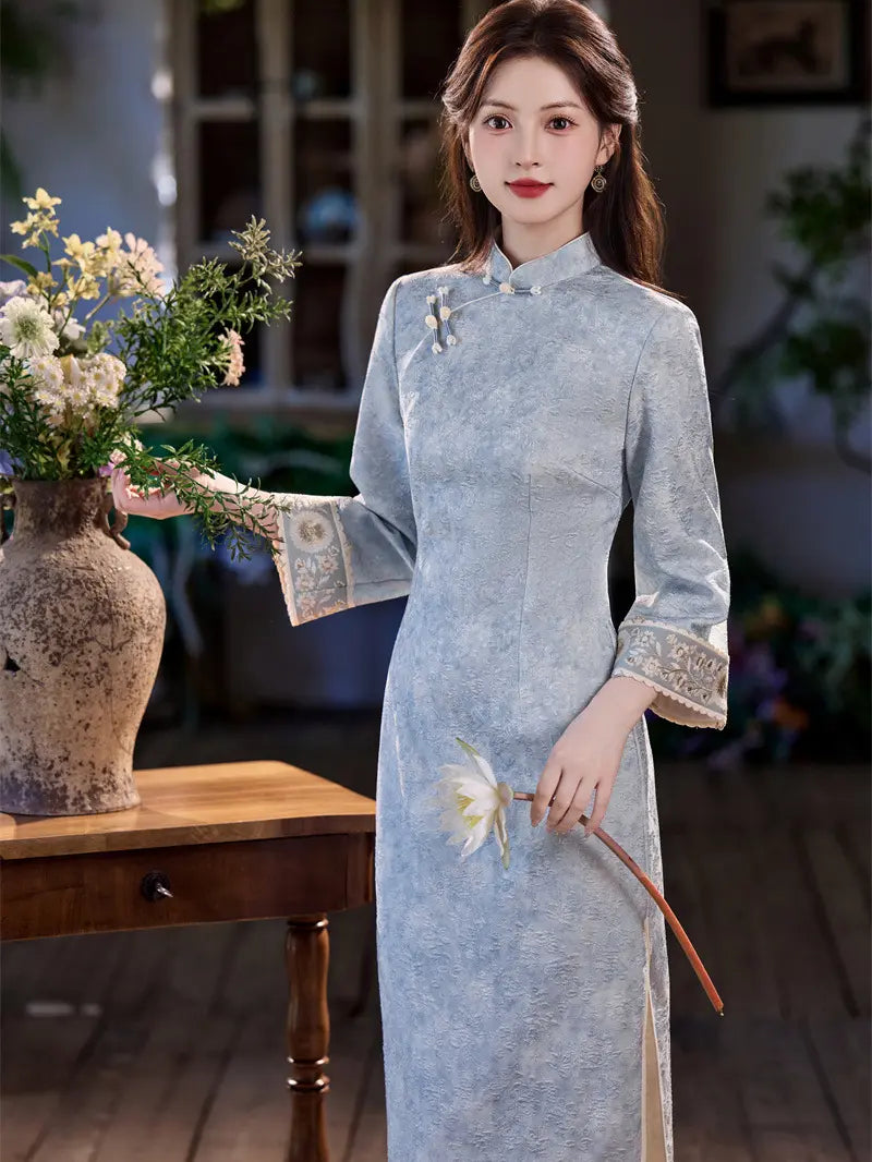 A woman wearing a light blue textured cheongsam (qipao) dress with embroidered cuffs and a floral brooch, standing in a rustic indoor setting next to a wooden table with a vase of flowers.