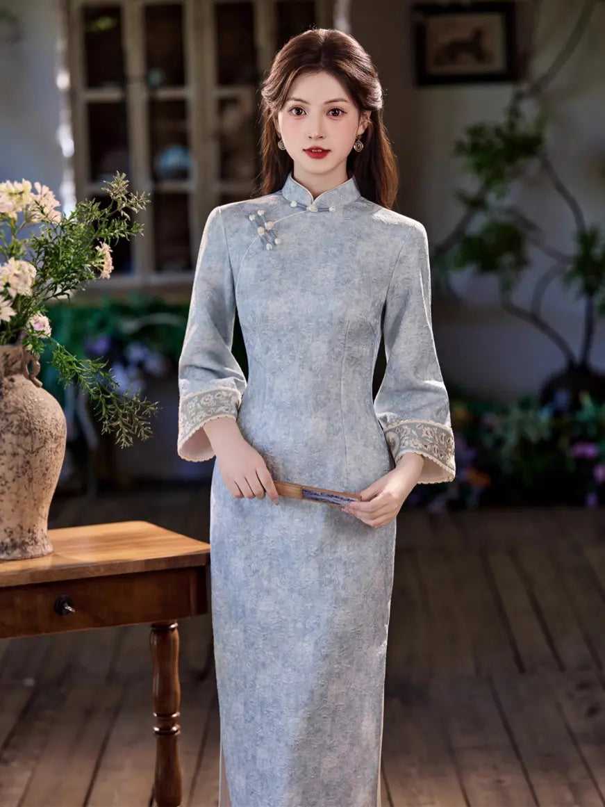 A woman stands indoors wearing a light blue-gray long cheongsam dress with lace trim at the cuffs and a small floral embellishment near the collar. She is posed beside a wooden table with flowers in a softly lit room with wood flooring and greenery in the background.