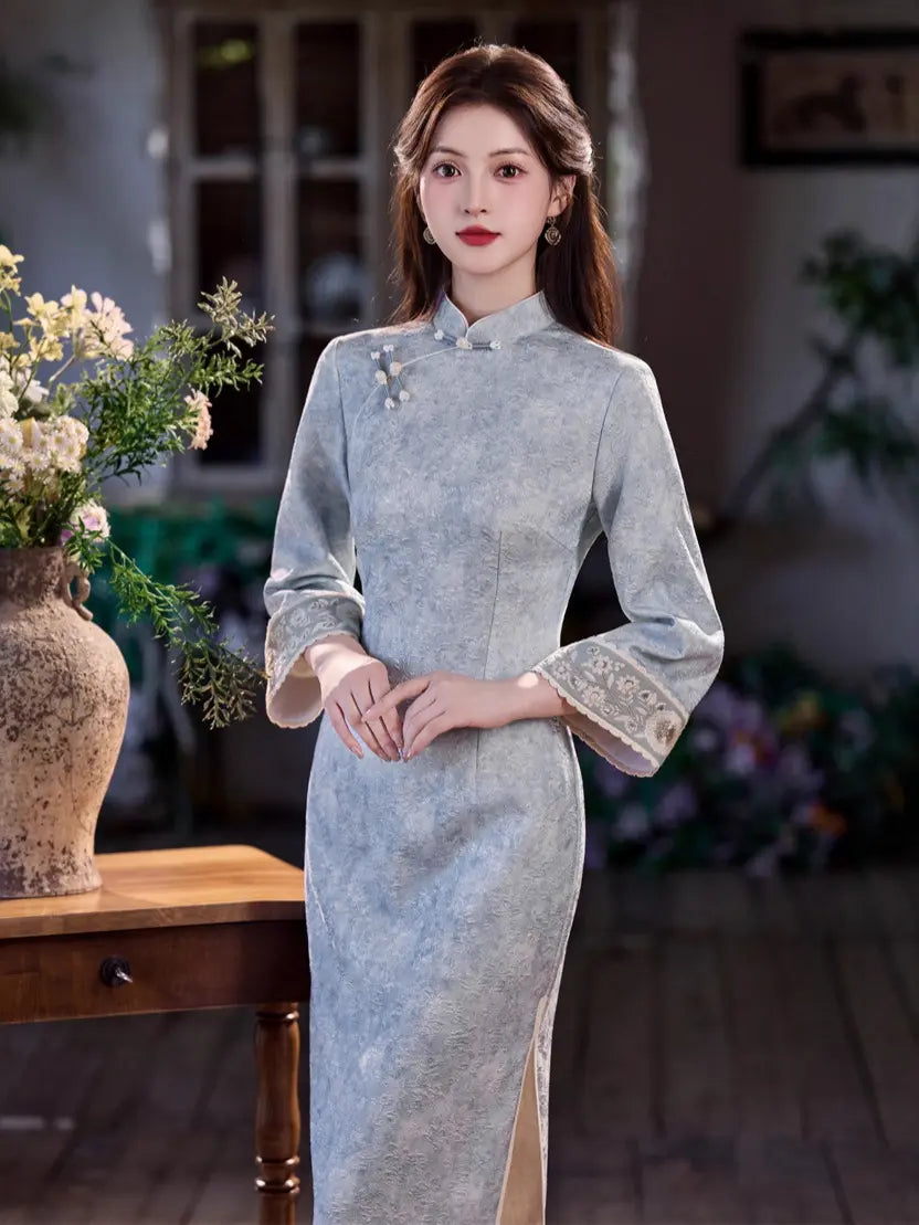 A woman wearing a light blue-gray traditional Chinese cheongsam dress with floral embroidery on the cuffs and collar, standing in a softly lit indoor setting with a wooden table and flowers in the foreground.