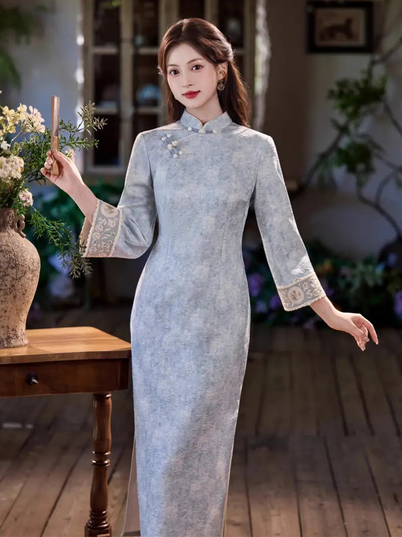 A woman wearing a light blue-gray textured cheongsam dress with embroidered cuffs and collar details, standing in a softly lit indoor setting with a wooden table and flowers beside her.