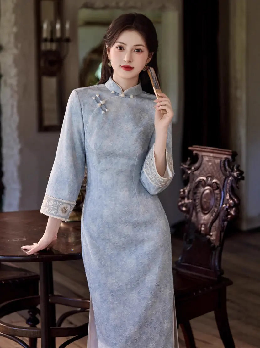 A woman wearing a light blue textured cheongsam (qipao) dress with lace trim on the cuffs and collar, standing in a vintage-style room next to a dark wooden table and chair.