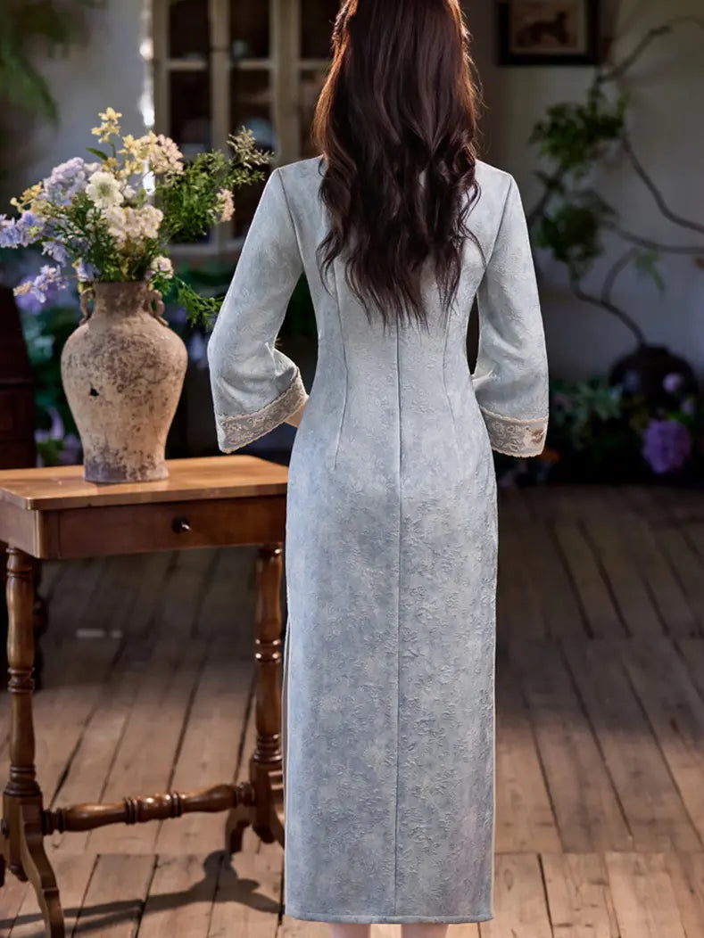 A woman viewed from behind wearing a long, light blue-gray textured dress with three-quarter sleeves and lace trim at the cuffs, standing in a rustic room with wooden floors and a table holding a vase of flowers.