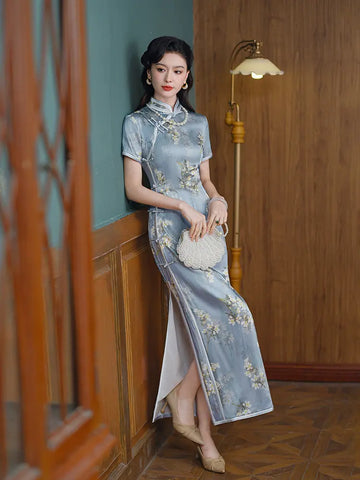 Woman in a Light Blue Mulberry Silk Floral Qipao Dressstanding in a room with wooden paneling and a lamp.