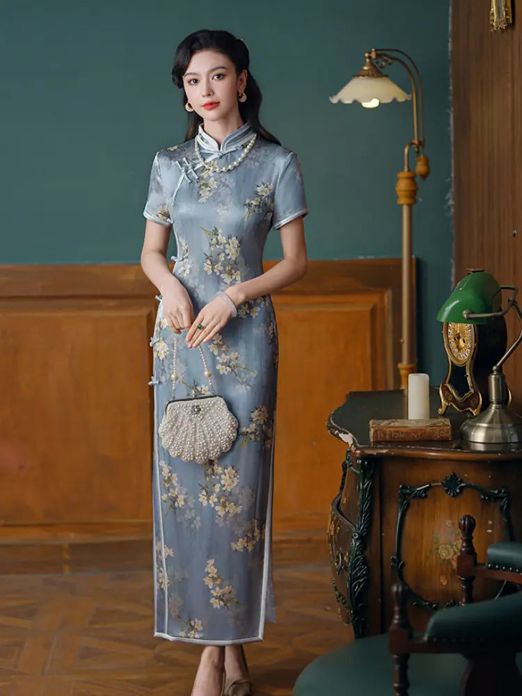 A woman stands indoors wearing a light blue Silk floral cheongsam/qipao dress with short sleeves, a high collar, and side slit hem. She is holding a small woven handbag and posed in a vintage room with wood paneling, a floor lamp, and an antique desk.