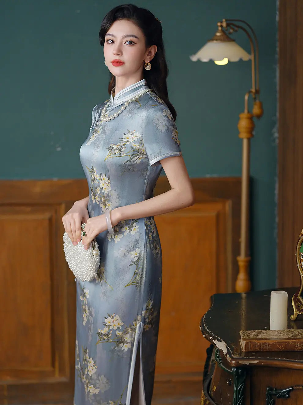 A woman wearing aLight Blue Mulberry Silk Floral Qipao  cheongsam dress, holding a white beaded clutch in a vintage-style room with a wooden floor and a lamp.