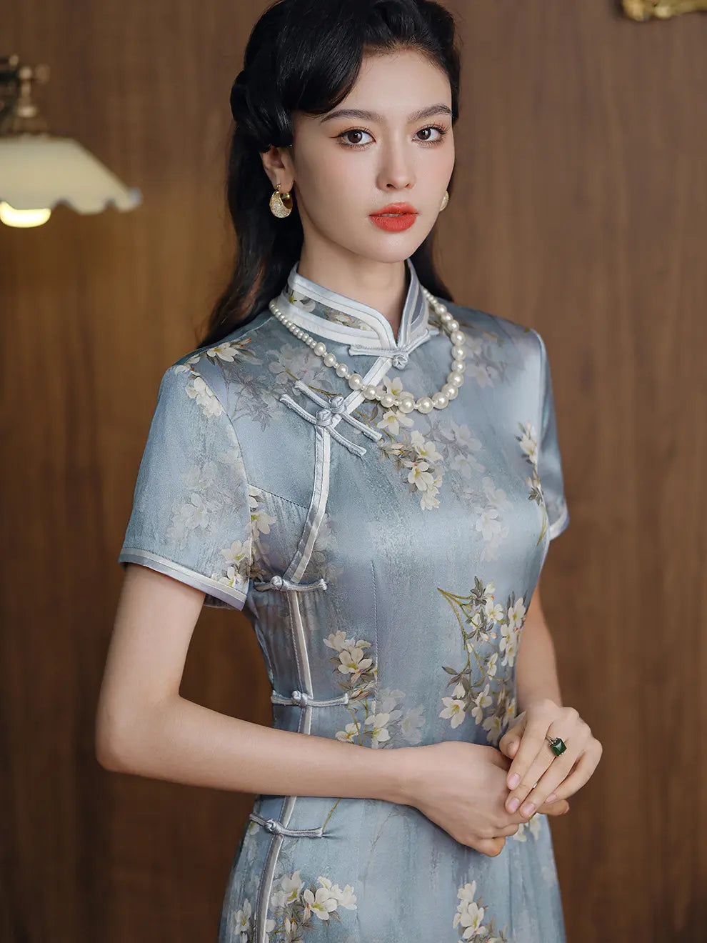 A woman wearing a light blue silk floral-printed cheongsam (qipao) dress with white trim and pearl necklaces, standing against a warm wood-paneled background.